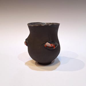 May include: A black ceramic vase with a unique design featuring red and white accents. The vase has a rounded shape and a slightly uneven rim.