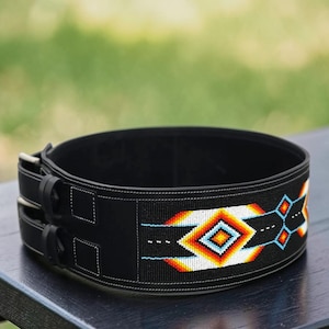 Puede incluir: Cinturón de halterofilia negro con un diseño decorativo de cuentas. El cinturón presenta un patrón geométrico en tonos naranja, blanco y azul. Tiene dos hebillas metálicas y está sobre una superficie de madera oscura.