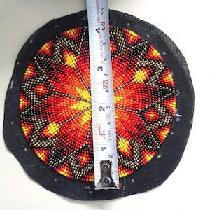 May include: A circular beaded design on a black fabric base. The design showcases a vibrant pattern of red, orange, yellow, and black beads. The diameter is approximately 7.6 cm. A tape measure is used to show the size.