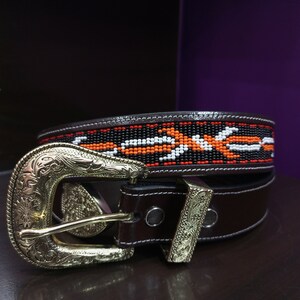 Puede incluir: Un cinturón de cuero marrón con una hebilla plateada y un diseño de cuentas en negro, blanco y naranja.