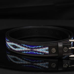 Op de afbeelding: Een zwarte leren riem met een centrale inleg van ingewikkeld kralenwerk in tinten blauw, paars en wit. De riem is voorzien van een zilverkleurige metalen gesp, bijpassende metalen klinknagels en witte contraststiksels langs de randen.