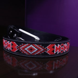 May include: Black leather belt with a beaded design in red, white, and black. The belt features a silver-tone buckle and a decorative pattern along the length. The belt is rolled up, showcasing the intricate beadwork.
