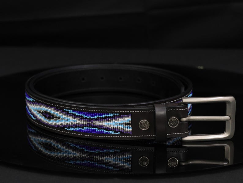 Op de afbeelding: Een zwarte leren riem met een centrale inleg van ingewikkeld kralenwerk in tinten blauw, paars en wit. De riem is voorzien van een zilverkleurige metalen gesp, bijpassende metalen klinknagels en witte contraststiksels langs de randen.