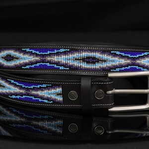 Op de afbeelding: Een zwarte leren riem met een centrale inleg van ingewikkeld kralenwerk in tinten koningsblauw, lichtblauw, wit en grijs. De riem is voorzien van een zilverkleurige metalen gesp, twee metalen klinknagels bij de gesp en witte contraststiksels.