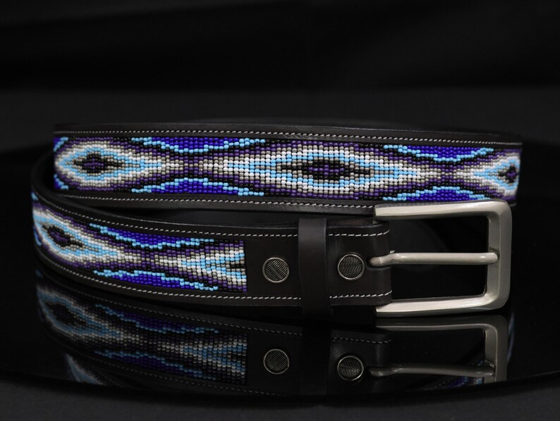 Op de afbeelding: Een zwarte leren riem met een centrale inleg van ingewikkeld kralenwerk in tinten koningsblauw, lichtblauw, wit en grijs. De riem is voorzien van een zilverkleurige metalen gesp, twee metalen klinknagels bij de gesp en witte contraststiksels.