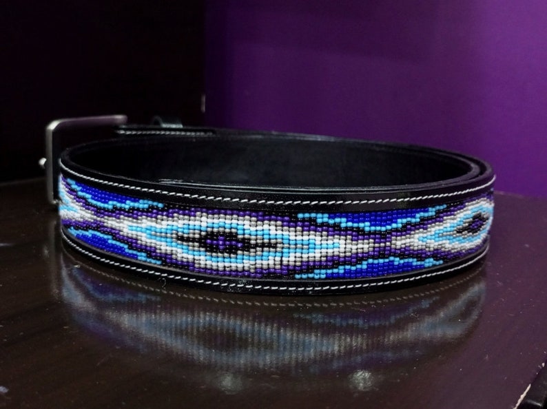 Op de afbeelding: Een zwarte leren riem met een centrale inleg van ingewikkeld kralenwerk in tinten koningsblauw, lichtblauw, paars, wit en zwart. De riem is voorzien van een zilverkleurige metalen gesp en witte stiksels langs de randen van de leren band.