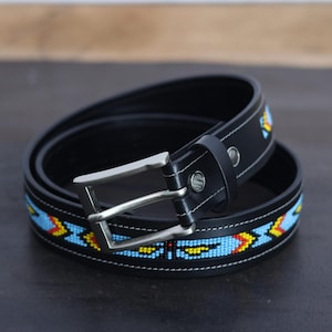Op de afbeelding: Een zwarte leren riem met een zilverkleurige metalen gesp en een decoratieve band van ingewikkeld kralenwerk. Het kralenwerk toont een geometrisch patroon in levendige tinten lichtblauw, geel en rood, gestikt langs het midden van de riem.