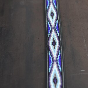 Op de afbeelding: Een lange, smalle riem met een centraal kralenpaneel met een herhalend ruitpatroon in tinten donkerblauw, lichtblauw, wit en zwart. Het kralengedeelte is omrand door donker materiaal met zichtbare witte stiksels langs de randen.