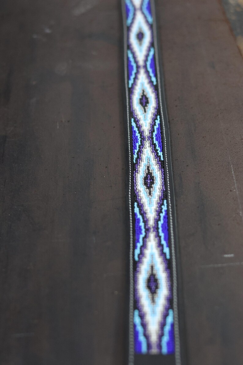 Op de afbeelding: Een lange, smalle riem met een centraal kralenpaneel met een herhalend ruitpatroon in tinten donkerblauw, lichtblauw, wit en zwart. Het kralengedeelte is omrand door donker materiaal met zichtbare witte stiksels langs de randen.