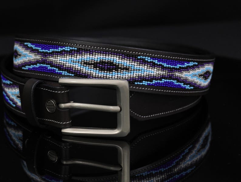 Op de afbeelding: Een zwarte leren riem met een centrale inleg van ingewikkeld kralenwerk in tinten koningsblauw, lichtblauw, wit en zwart. De riem is voorzien van een matzilverkleurige metalen gesp en een enkele leren lus, met zichtbare witte stiksels.