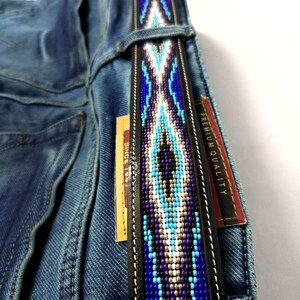 Op de afbeelding: Een zwarte leren riem met een centrale strook van ingewikkeld kralenwerk in tinten blauw, paars, wit en zilver, door de riemlussen van een blauwe spijkerbroek gehaald, met een label met de tekst PREMIUM QUALITY zichtbaar aan de zijkant.
