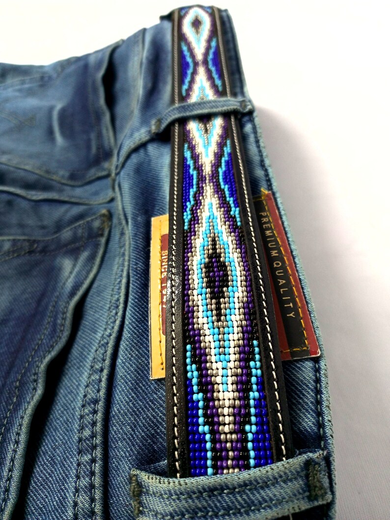 Op de afbeelding: Een zwarte leren riem met een centrale strook van ingewikkeld kralenwerk in tinten blauw, paars, wit en zilver, door de riemlussen van een blauwe spijkerbroek gehaald, met een label met de tekst PREMIUM QUALITY zichtbaar aan de zijkant.