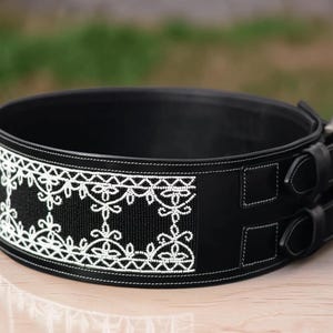 May include: A black weightlifting belt with white decorative embroidery. The belt features a detailed floral pattern and two buckle closures. The belt is designed for support during weightlifting exercises.