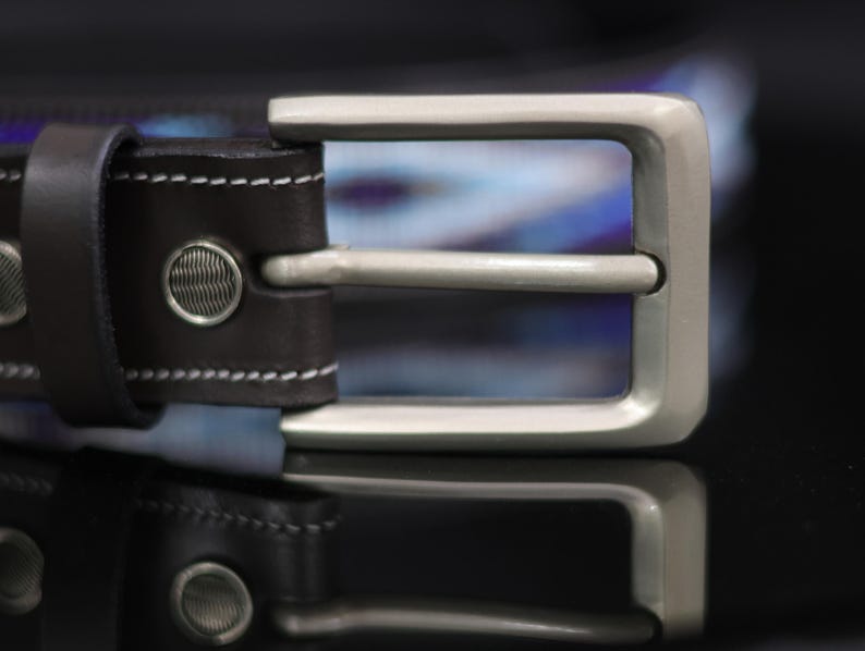 Op de afbeelding: Een donkerbruine leren riem met een rechthoekige gesp van geborsteld zilverkleurig metaal en een bijpassende leren lus. De riem is afgewerkt met witte contraststiksels en twee ronde zilveren klinknagels met een textuurpatroon.
