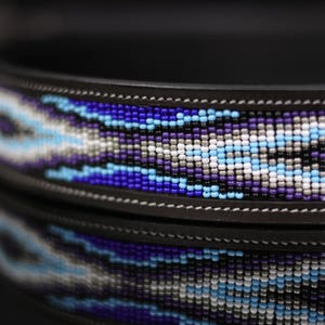 Op de afbeelding: Een donkerbruine leren halsband met een centrale inleg van complexe kraaltjes in de kleuren koningsblauw, lichtblauw, paars, wit en zwart, gerangschikt in een herhalend geometrisch patroon met zichtbare witte stiksels langs de randen.
