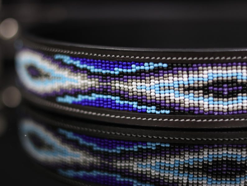 Op de afbeelding: Een donkerbruine leren halsband met een centrale inleg van complexe kraaltjes in de kleuren koningsblauw, lichtblauw, paars, wit en zwart, gerangschikt in een herhalend geometrisch patroon met zichtbare witte stiksels langs de randen.