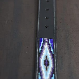 Op de afbeelding: Een zwarte leren riem met een centrale inleg van ingewikkeld kralenwerk in tinten paars, lichtblauw, wit en zwart. Het kralenwerk vormt een herhalend ruitpatroon langs de riem, die is voorzien van witte stiksels en metalen gaatjes.