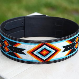 Può includere: Una cintura in pelle nera con un design di perline. Il motivo a perline presenta forme geometriche in bianco, nero, arancione e turchese. La cintura ha una chiusura con fibbia.