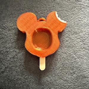 Può includere: Ghiacciolo arancione a forma di Topolino stampato in 3D con un morso sull'orecchio. Il ghiacciolo ha un centro cavo e un bastoncino di legno. L'articolo è su una superficie grigia strutturata.