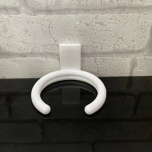 May include: White plastic wall-mounted holder for a coffee mug or cup. The holder is shaped like a ring and has a flat back.