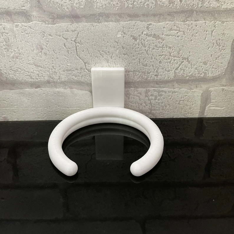 May include: White plastic wall-mounted holder for a coffee mug or cup. The holder is shaped like a ring and has a flat back.