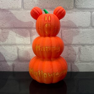 Puede incluir: Una pila de tres calabazas naranjas decoradas con una cara de Mickey Mouse y las palabras "Happy Halloween".