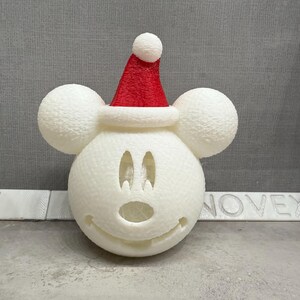 May include: White styrofoam Mickey Mouse head ornament with a red Santa hat. The ornament has a cut-out face with black eyes and a nose.