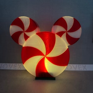 Puede incluir: Una luz con forma de caramelo de menta roja y blanca con una base verde. La luz tiene forma de cabeza de Mickey Mouse.