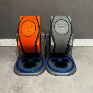May include: Two Disney MagicBands, one orange and one gray, are displayed in a 3D printed stand. Each stand features a blue Mickey Mouse silhouette.