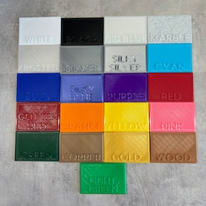 May include: A collection of 16 small squares, each showing a different color and texture of plastic filament. The colors include white, black, blue, gray, silver, cyan, purple, red, orange, yellow, pink, green, copper, gold, wood, and light green.