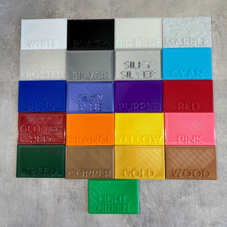 May include: A collection of 16 small squares, each showing a different color and texture of plastic filament. The colors include white, black, blue, gray, silver, cyan, purple, red, orange, yellow, pink, green, copper, gold, wood, and light green.