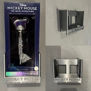 May include: A silver key-shaped ornament with a Mickey Mouse head and Space Mountain design. The ornament is in a blue box with the words "Mickey Mouse The Main Attraction" on the front. The box is sitting on a silver stand with the same words on it.