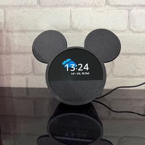 May include: Black smart speaker with a circular screen showing the time 13:24 and the date. The speaker has a Mickey Mouse ear design. The speaker is on a black surface.
