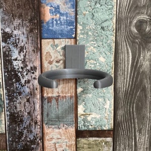 May include: A gray metal cup holder mounted on a wood panel wall. The holder is shaped like a half-circle and has a smooth, rounded edge.