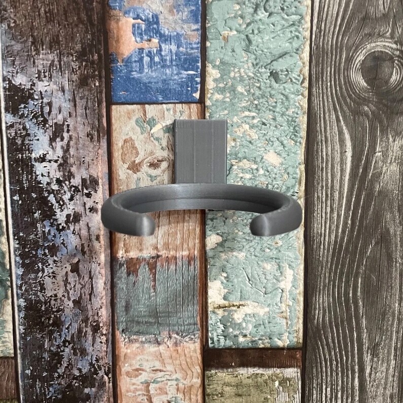 May include: A gray metal cup holder mounted on a wood panel wall. The holder is shaped like a half-circle and has a smooth, rounded edge.
