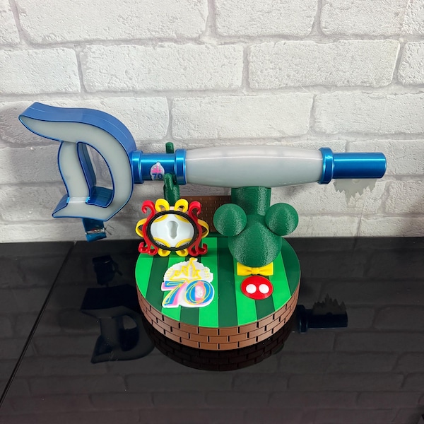 May include: A whimsical, 3D-printed wand with a blue and white design, featuring a Mickey Mouse-themed base with a green field, red and yellow accents, and the number "70". The wand has a blue handle and a white and blue tip.