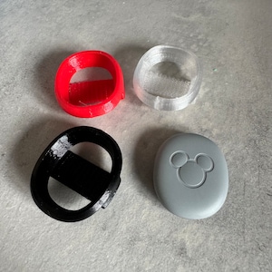 Può includere: Quattro oggetti diversi stampati in 3D. Un oggetto rosso, un oggetto nero, un oggetto trasparente e un oggetto grigio. L'oggetto grigio ha una silhouette di Topolino.