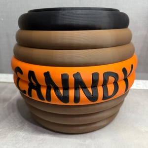 May include: A 3D-printed candy container with a stacked, cylindrical design. The container features a black top, a brown midsection, and an orange band with the word "CANDY" in black lettering. The base is a darker brown.