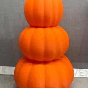 Può includere: Una pila di tre zucche arancioni con steli verdi. Le zucche sono di un arancione brillante e presentano scanalature verticali. Le zucche sono impilate, creando un'esposizione decorativa autunnale.
