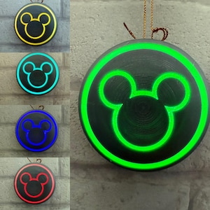 May include: A black circular ornament with a Mickey Mouse silhouette design. The ornament has a glowing outline and the Mickey Mouse design is illuminated in yellow, blue, green, cyan, and red. A gold string is attached for hanging.