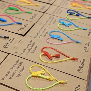 Op de afbeelding: Een set bruine kaarten met kleurrijke rits-hartbedels eraan bevestigd. Elke kaart heeft een boodschap erop geschreven, waaronder "You couldn't be any hipper - so here's a friendship fidget zipper!" en "Melita".