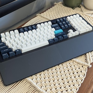 Puede incluir: Un teclado mecánico blanco y negro con un juego de teclas azul y blanco. El teclado tiene un reposamuñecas grande y un teclado numérico dedicado.