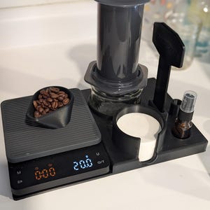 以下が含まれることがあります： 黒いデジタルスケールに、灰色のコーヒーカップホルダーと灰色のエアロプレスコーヒーメーカーが置かれています。スケールには「0:00」と「20.0」が表示されています。小さな黒いスプーンにコーヒー豆が入っています。