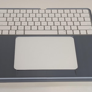 Puede incluir: Un combo de teclado y trackpad inalámbrico plateado y blanco. El teclado tiene teclas blancas con letras negras. El trackpad es una superficie blanca lisa. El dispositivo está enmarcado en un marco gris oscuro.