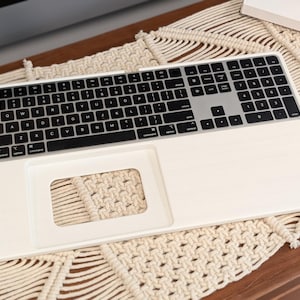 Puede incluir: Un teclado blanco con teclas negras y un área de panel táctil plateada. El teclado tiene un recorte rectangular para un panel táctil. El teclado está sobre una superficie de macramé.