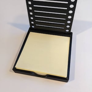 May include: Black 3D printed desk organiser with a slot for sticky notes and a side compartment with holes for pens or pencils.