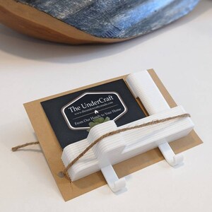 Peut inclure: Porte-manteau en plastique blanc avec quatre crochets, attaché avec de la ficelle. Le porte-manteau est sur une carte brune avec le texte "The UnderCraft" et "From Our Hands to Your Home".