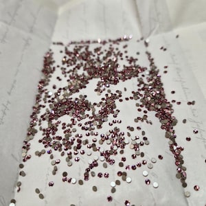 Könnte beinhalten: Eine Streuung kleiner, runder, rosa und silberner Strasssteine auf einer weißen Papieroberfläche. Die Strasssteine sind unterschiedlich groß und reflektieren das Licht, wodurch ein funkelnder Effekt entsteht. Das Papier hat eine schwache, dekorative Schrift.