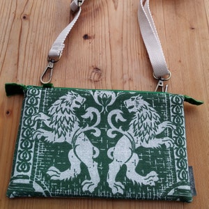May include: A green fabric zippered pouch with a white strap. The pouch features a white design of two lions facing each other.