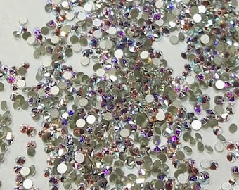 May include: A large quantity of iridescent clear glass rhinestones, also known as crystals, scattered on a white surface. The rhinestones have a flat back with a silver-colored adhesive backing.